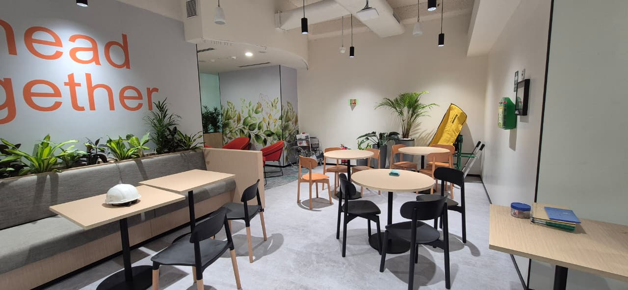 GSK office breakout lounge with the 'Ahead Together' feature wall, planted bench and café tables, delivered by Ananya Interiors in Bengaluru