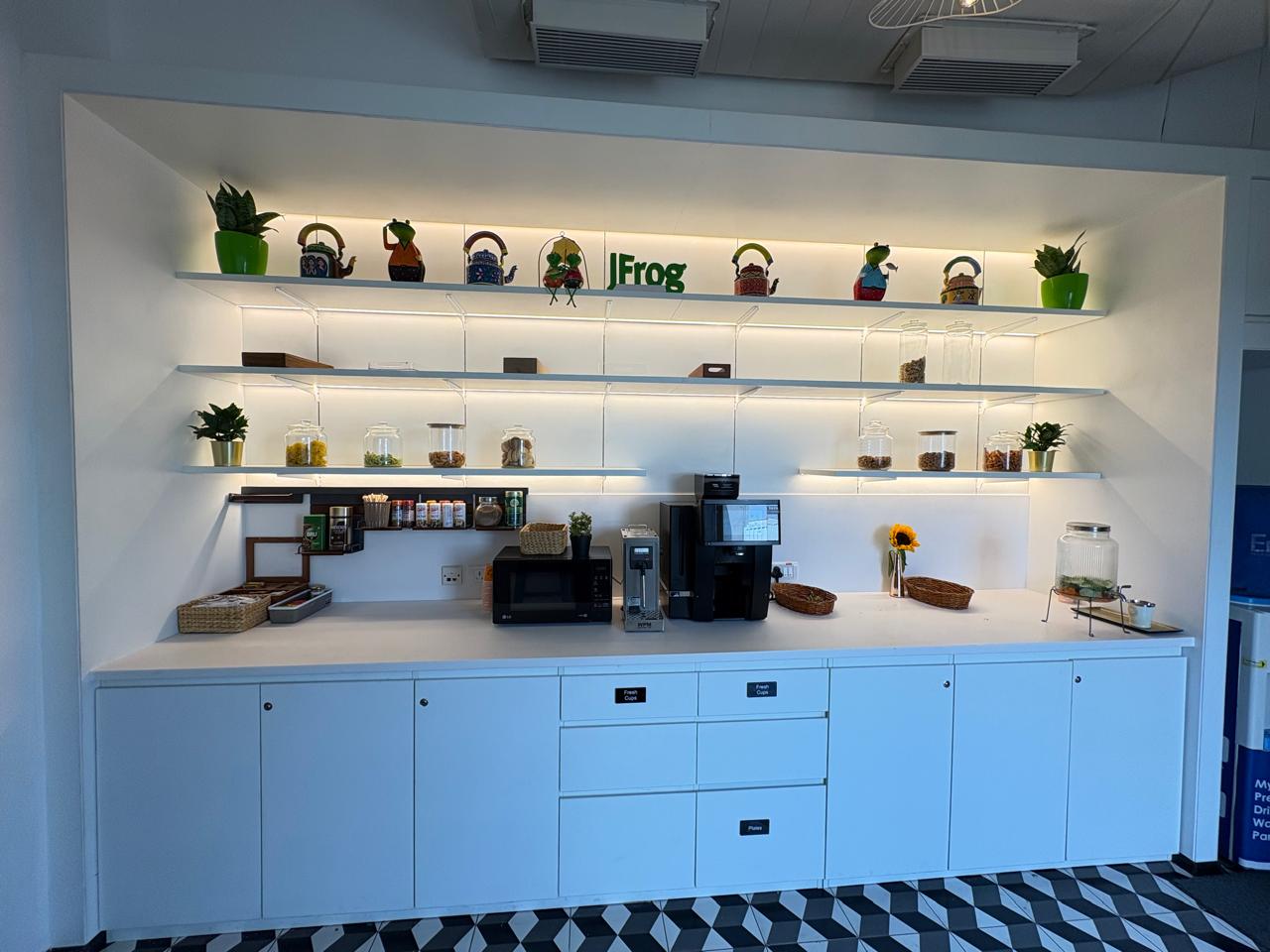 JFrog India office pantry wall with LED-lit shelves and the green JFrog logo above the tea point, delivered by Ananya Interiors in Bengaluru